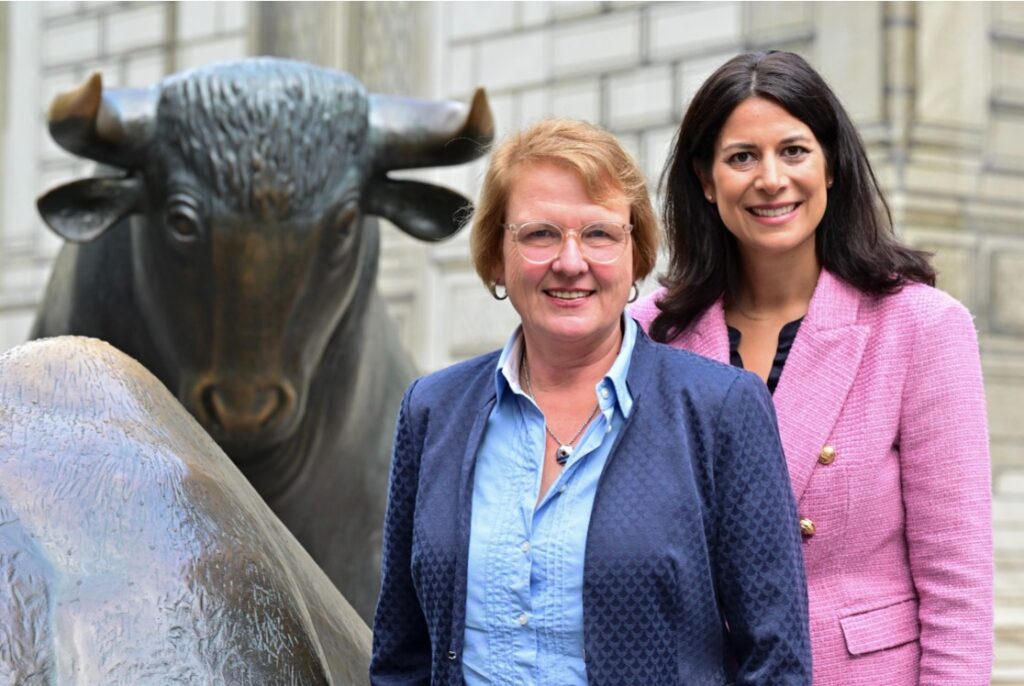 Zwei lächelnde Frauen vor einer bronzenen Bullenstatue, eine im blauen, die andere im rosa Blazer.
