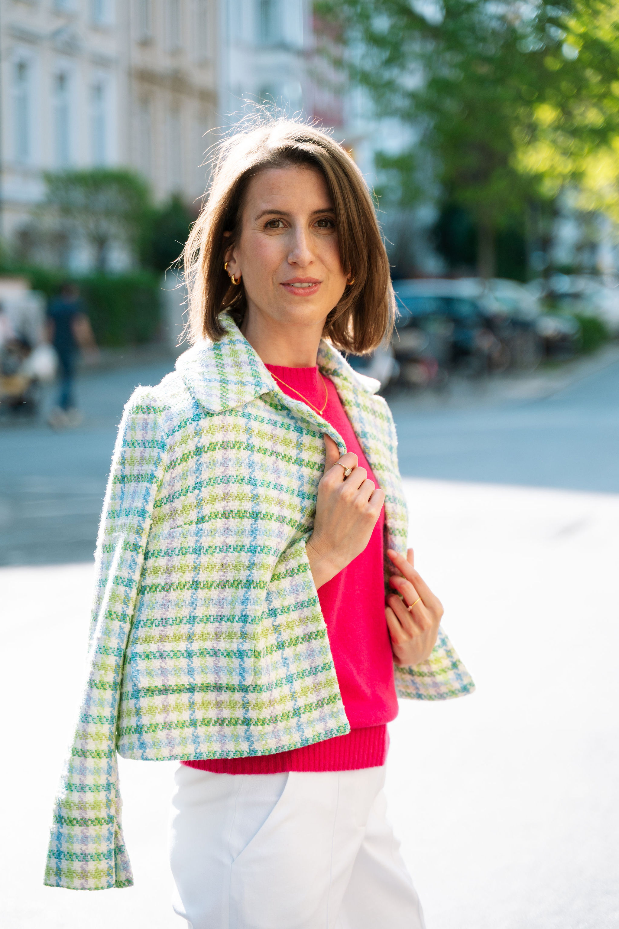 Eine Frau lächelt, trägt eine bunte Jacke und ein pinkes Oberteil, steht draußen bei sonnigem Wetter.