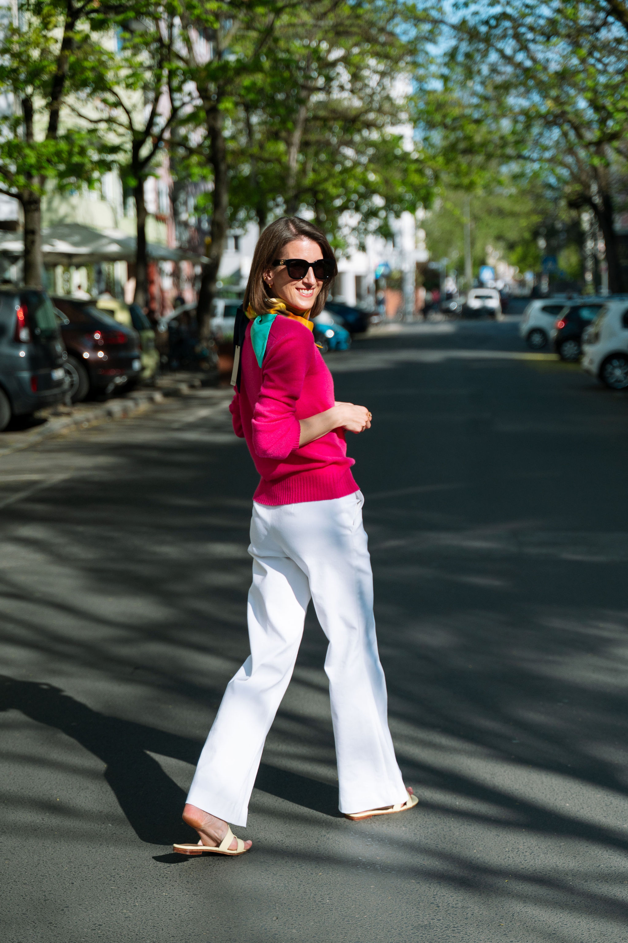 Eine Frau lächelt auf einer sonnigen Straße, sie trägt rosa Oberteil, weiße Hose, Sonnenbrille.