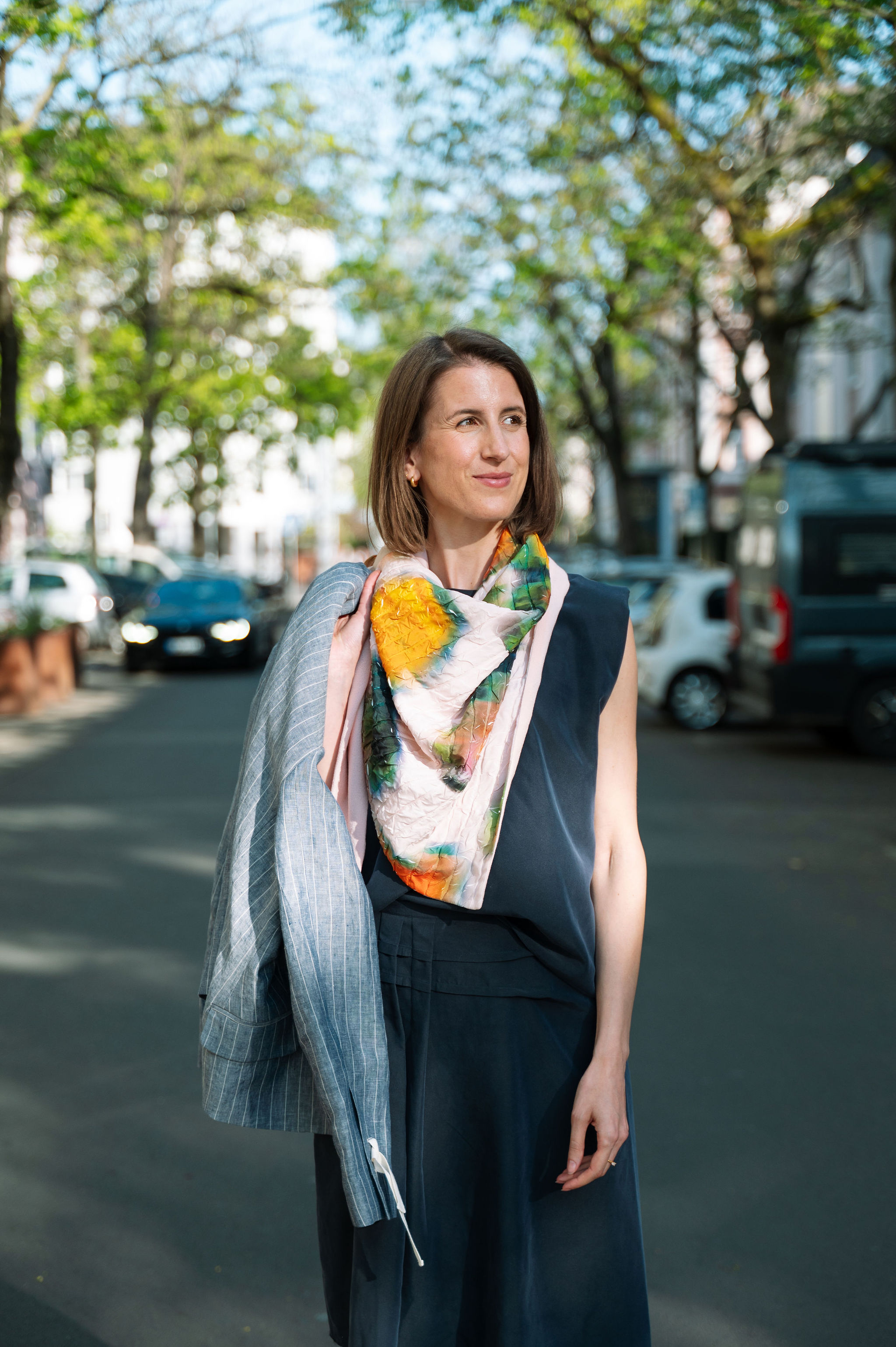 Frau in blauem Kleid, gestreiftem Mantel, buntem Schal, lächelnd, steht auf sonniger Straße.
