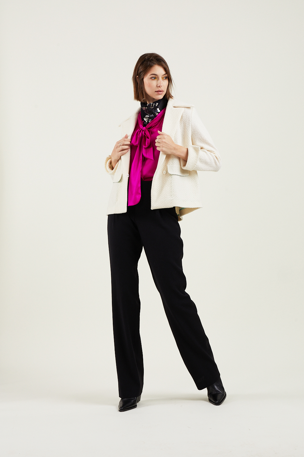 Frau mit ernstem Blick in weißem Blazer, rosa Bluse und schwarzer Hose auf cremefarbenem Hintergrund.