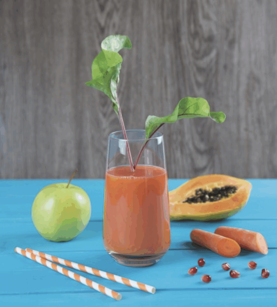 Ein Glas mit orangem Saft, Garnitur, Apfel, Papaya, Karotte und Strohhalme auf blauem Tisch.