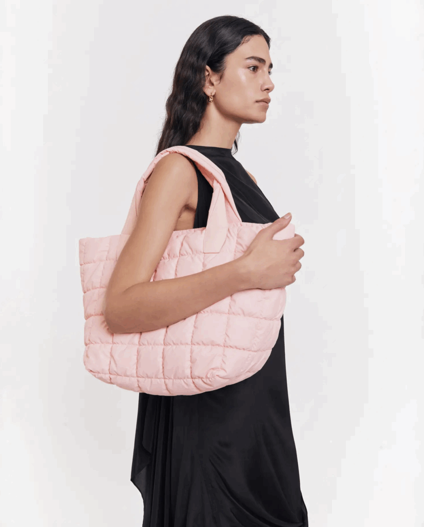 Eine Frau im Profil, neutrale Miene, schwarze Kleidung, rosa gesteppte Tasche über der Schulter.
