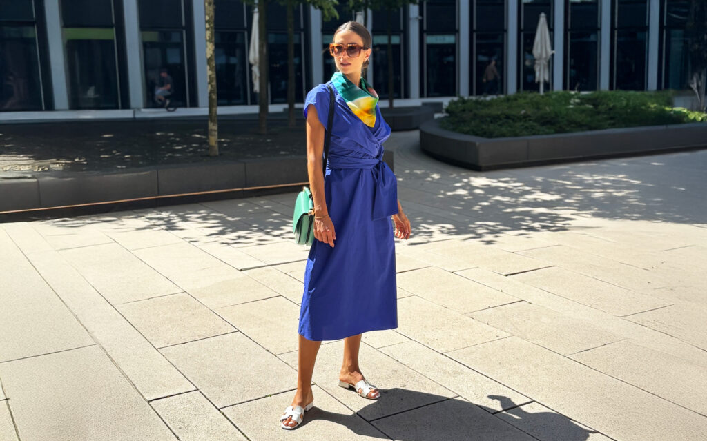 Eine Frau im blauen Kleid steht draußen, trägt eine Sonnenbrille und ist elegant gekleidet