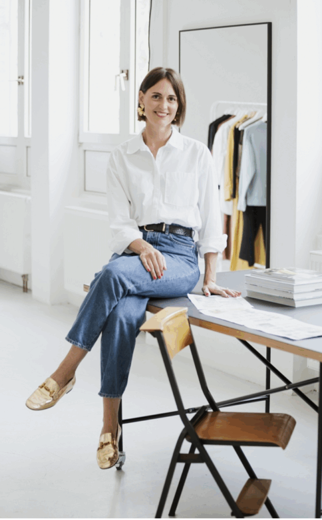 Eine lächelnde Frau sitzt lässig auf einem Tisch in einem hellen Raum mit Spiegel.